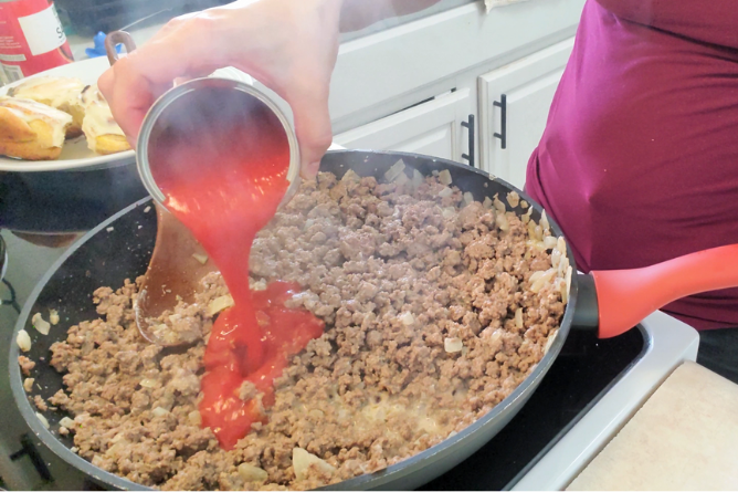 woman pouring a can of tomato sauce into finely chopped hamburger meat