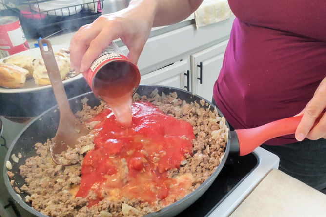 woman pouring water from a can of tomato sauce into a skillet full of hamburger meat