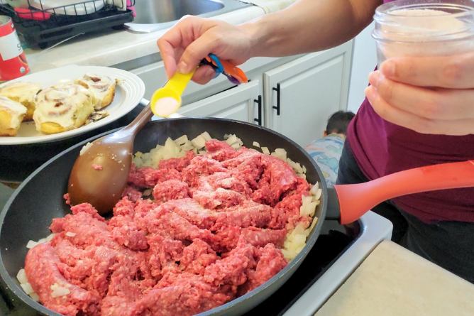 woman adding a teaspoon of salt into a skillet filled with hamburger meat and onions